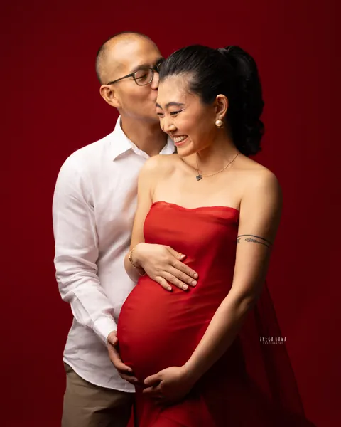 Pregnancy photoshoot in Gurgaon: husband in a white shirt kissing his laughing wife's cheek, she wears a strapless red drape and holds her