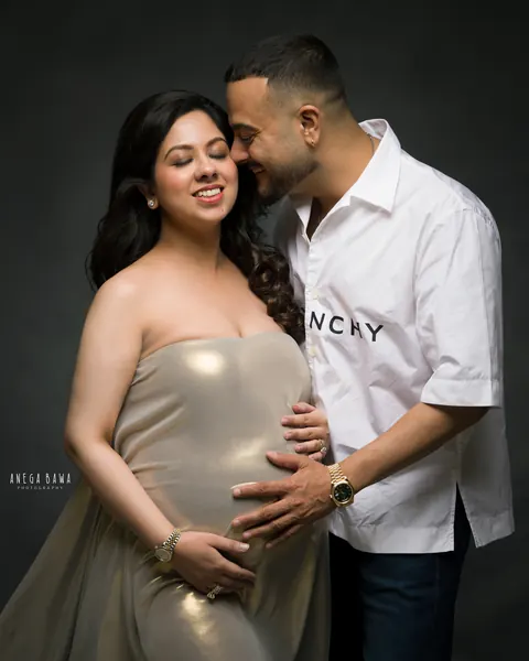 Pregnancy photoshoot in Gurgaon: smiling expecting mother in a strapless gold metallic drape while her husband in a white shirt nuzzles her