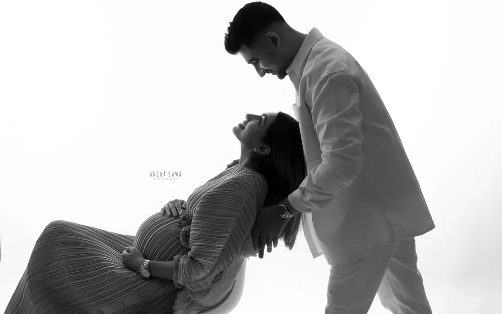 Pregnancy photoshoot in Gurgaon: backlit black and white portrait of a husband leaning over his wife who reclines on a white chair in a