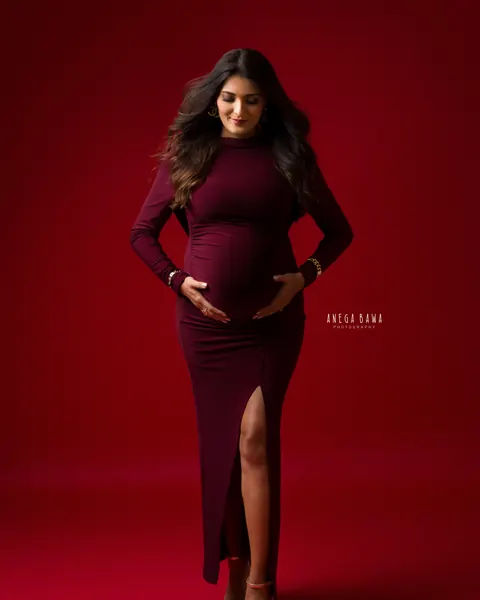 Pregnancy photoshoot in Gurgaon: expecting mother walking toward the camera in a maroon gown with a side slit and gold heels, looking down