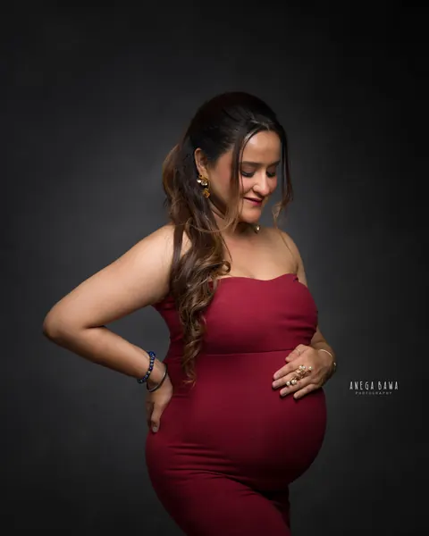 Pregnancy photoshoot in Gurgaon: expecting mother in a maroon strapless gown with one hand on her hip and one on her bump, looking down