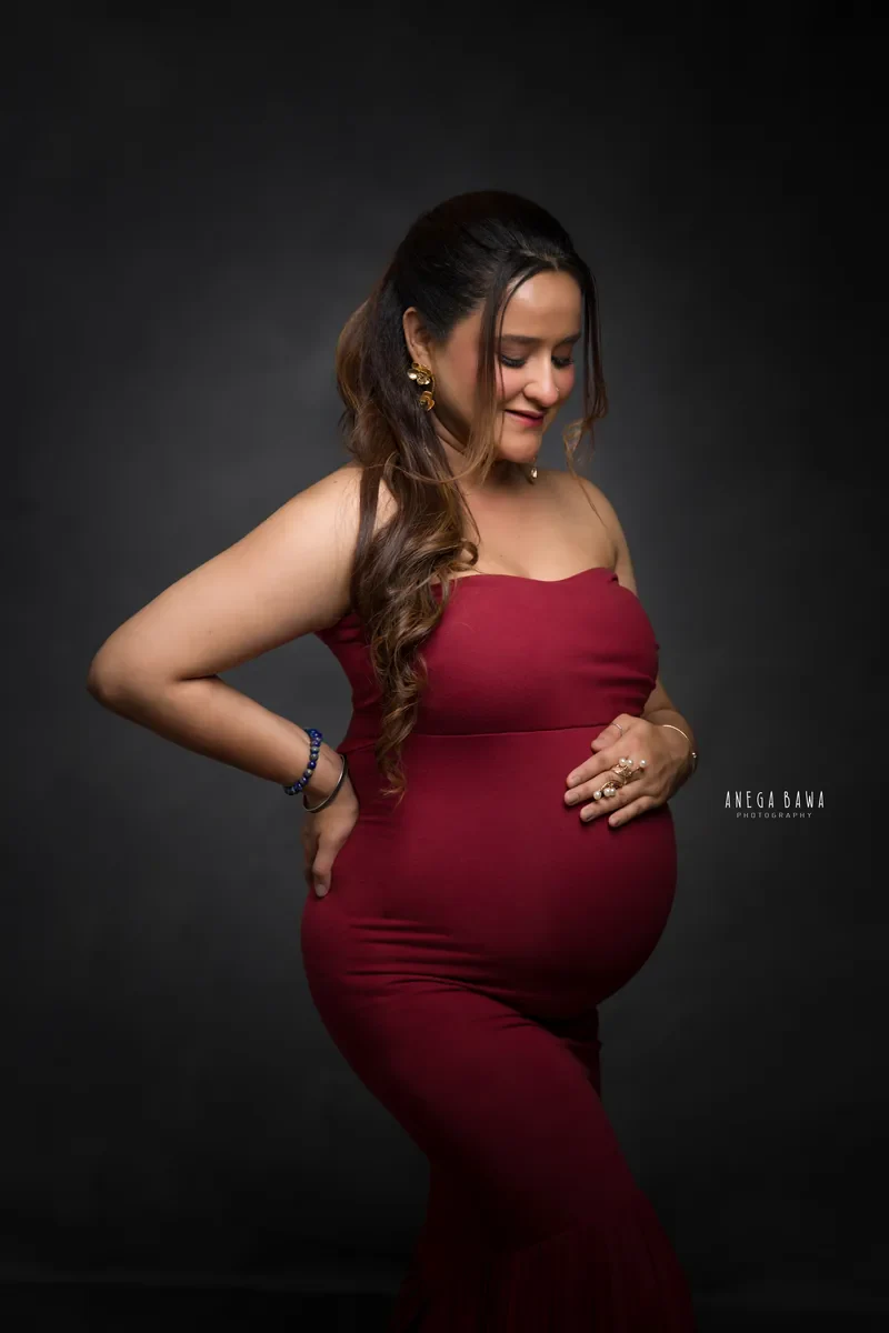 Pregnancy photoshoot in Gurgaon: expecting mother in a maroon strapless gown with one hand on her hip and one on her bump, looking down