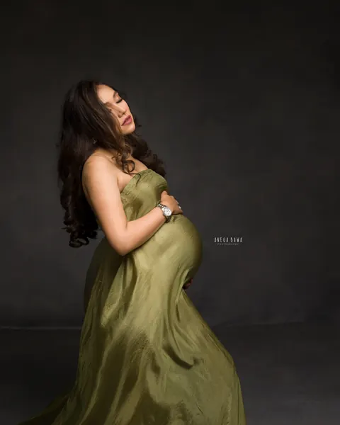 Pregnancy photoshoot in Gurgaon: side profile of expecting mother in an olive green satin drape, head tilted back, dark backdrop, by Anega