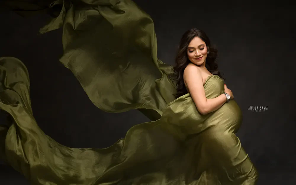 Pregnancy photoshoot in Gurgaon: expecting mother smiling down at her bump in a strapless olive green drape with fabric swirling around