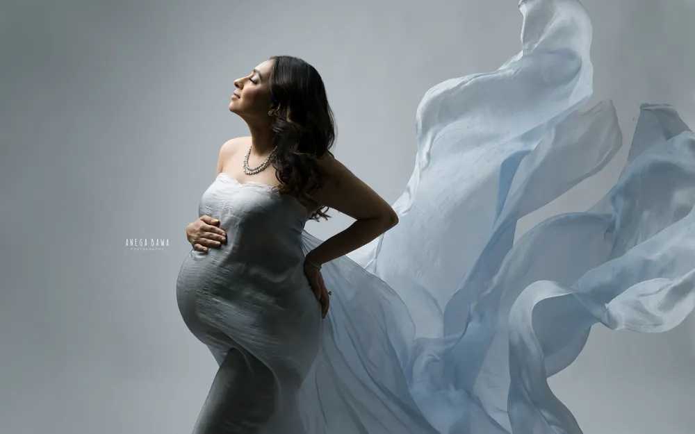 Pregnancy photoshoot in Gurgaon: expecting mother in profile with her face lifted, wearing a powder blue silk drape that billows behind