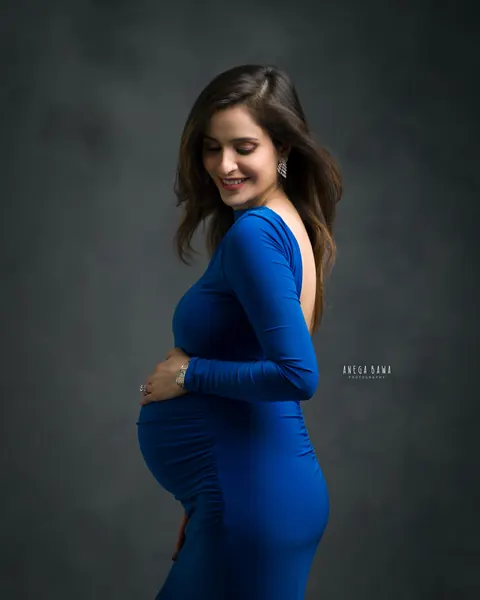 Pregnancy photoshoot in Gurgaon: expecting mother in a royal blue open-back gown looking down over her shoulder with a hand on her bump