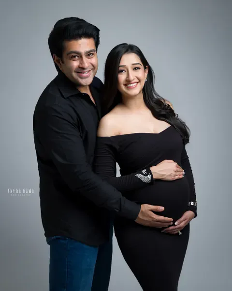 Pregnancy photoshoot in Gurgaon: smiling couple hugging, wife in a black off-shoulder gown with embroidered cuff and husband in a black