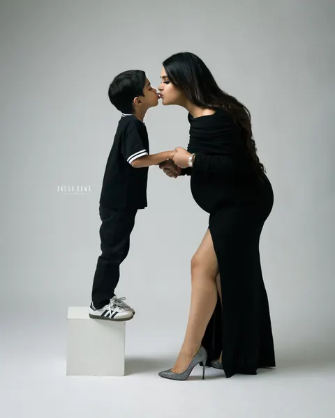 Pregnancy photoshoot in Gurgaon: young son standing on a white block to kiss his expecting mother, who wears a black off-shoulder gown with