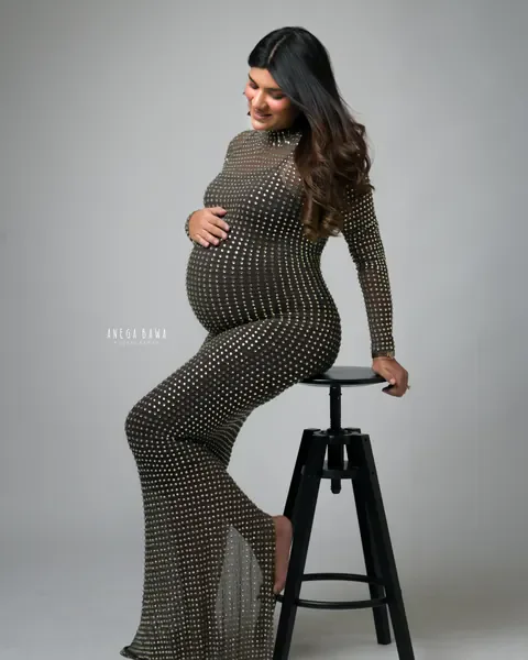Pregnancy photoshoot in Gurgaon: expecting mother seated on a black stool in a fitted crystal-studded sheer gown, hand on her bump, grey