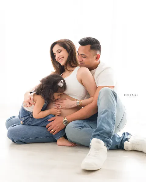 Pregnancy photoshoot in Gurgaon: toddler hugging her mother's bump while her father holds them both, family seated on a white floor in