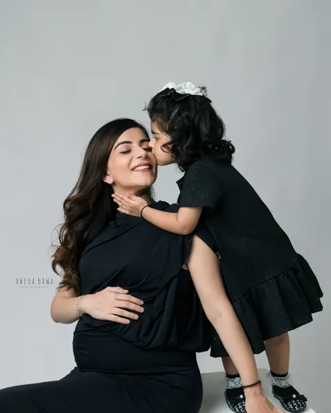 Pregnancy photoshoot in Gurgaon: toddler kissing her expecting mother's cheek, mother in a black gown seated on a white block, grey