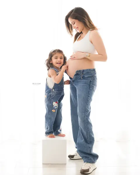 Pregnancy photoshoot in Gurgaon: toddler in denim dungarees standing on a white block and touching her mother's bare bump, mother in a