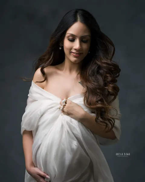 Pregnancy photoshoot in Gurgaon: smiling expecting mother in a white silk drape off the shoulders with long wavy hair and pearl rings, one