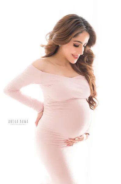 Smiling expecting mother in a blush pink off-shoulder long-sleeve fitted dress, wavy hair, hands on her bump and hip, bright white backdrop.