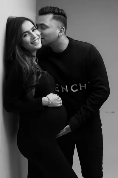 Black and white couple portrait: smiling wife in a black fitted dress leaning on a wall as her husband in a black sweatshirt kisses her