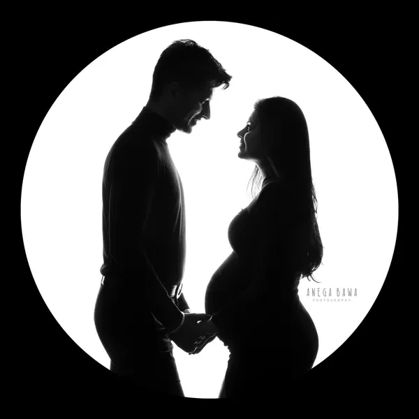 Silhouette of a couple facing each other holding hands over the bump, husband in a dark top, wife in profile, inside a white circle on a