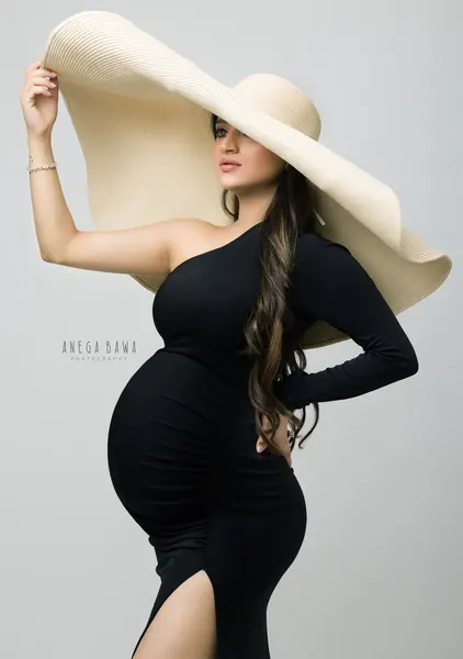 Expecting mother in a fitted black one-shoulder long-sleeve dress with a high slit and an oversized cream straw hat, hand on hip, light