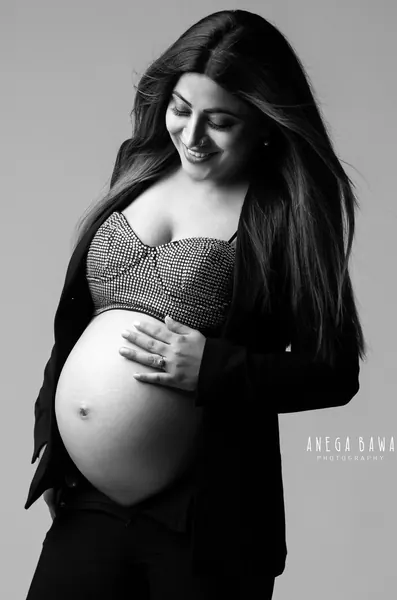 Black and white portrait of smiling expecting mother in an open black blazer and studded bralette, long straight hair, bare bump, hand on