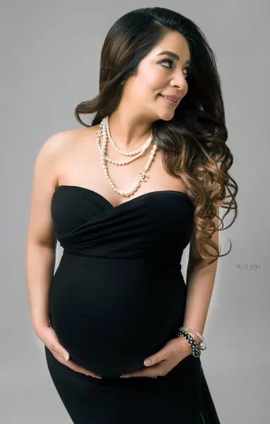 Smiling expecting mother in a black strapless sweetheart-neck gown with layered pearl necklaces and beaded bracelets, looking to the side
