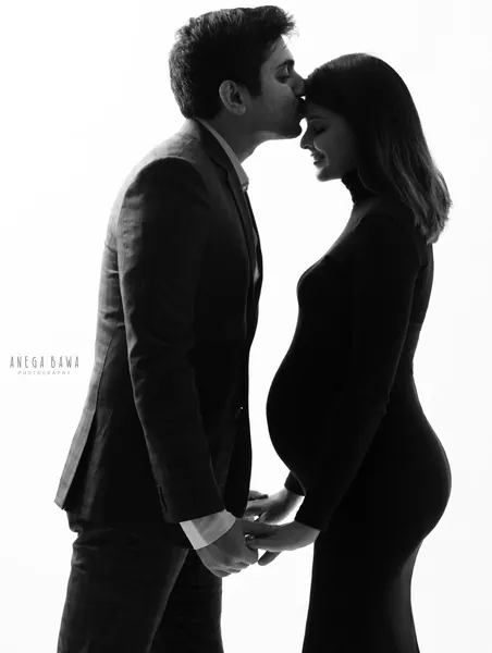 High-key black and white silhouette of a couple, husband in a blazer kissing his wife's forehead while holding hands, wife in a black
