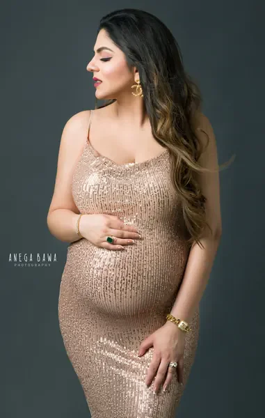 Expecting mother in a champagne sequin strap dress with gold earrings, long ombre hair, looking to the side, hand on her bump, dark teal