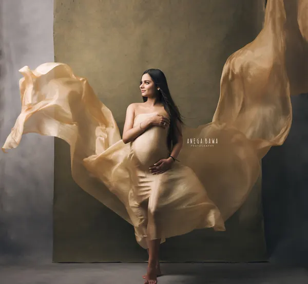 Expecting mother in a champagne gold silk drape with large sheer wings of fabric flying out on both sides, barefoot, hand on her bump