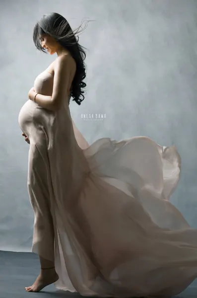 Expecting mother in profile in a pearl-grey satin drape with a long flowing train, black hair blowing across her face, barefoot, grey-blue