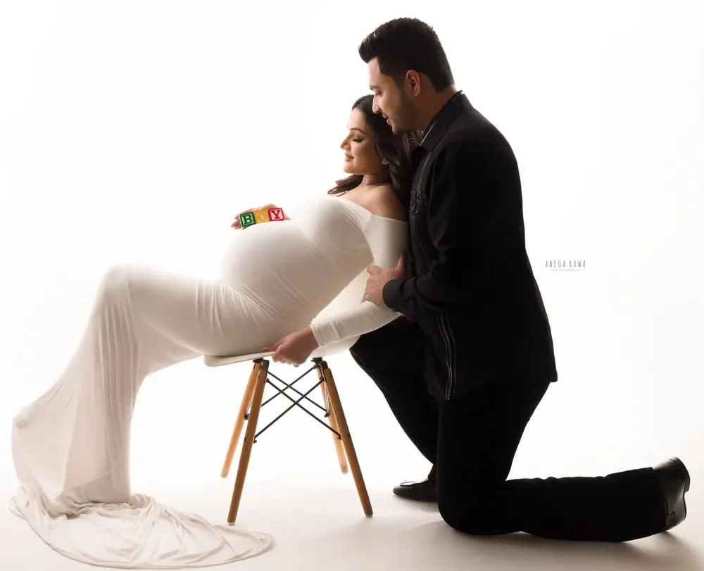 Expecting mother in a white off-shoulder fitted gown reclining on a wooden chair with alphabet blocks on her bump, husband in a black