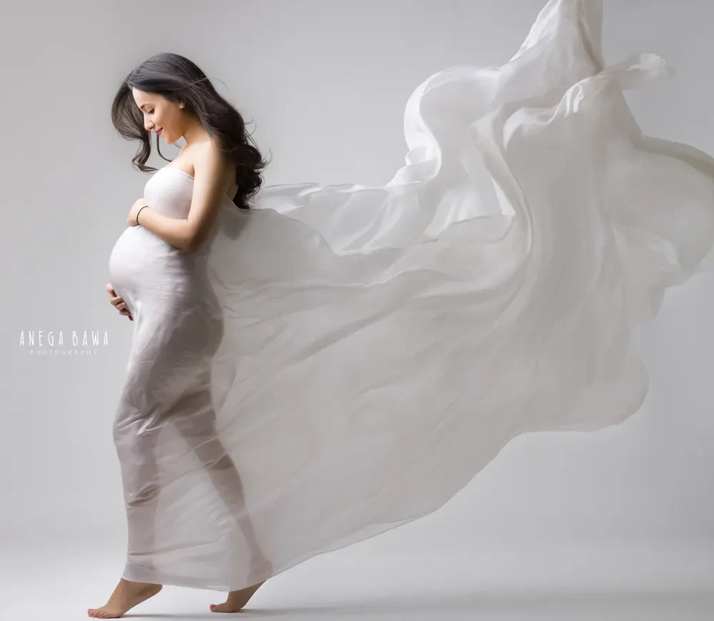 Expecting mother in a white fitted strapless gown with a large sheer white drape flying out behind her, barefoot, looking down at her bump