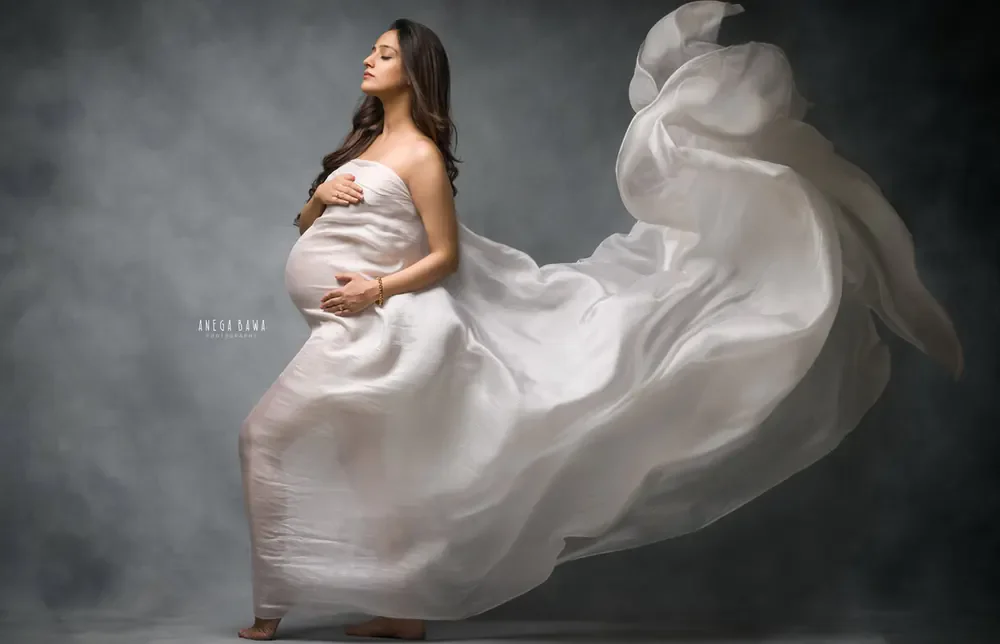 Expecting mother in a sheer white drape with a huge billowing train flying behind her, hands on her bump, looking up, dark grey vignette