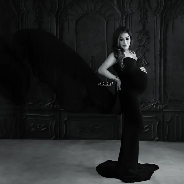 Black and white portrait of expecting mother in a black strapless fitted gown with a train, a black sheer drape flying beside her, hand on