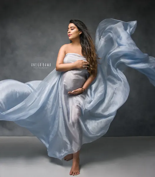Expecting mother in a pale blue sheer satin drape wrapped across her chest with fabric flying out in a huge swirl, barefoot, hand on her