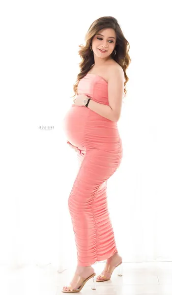 Expecting mother in a coral pink ruched strapless midi dress and clear heels, hands on her bump, looking to the side, bright white backdrop.