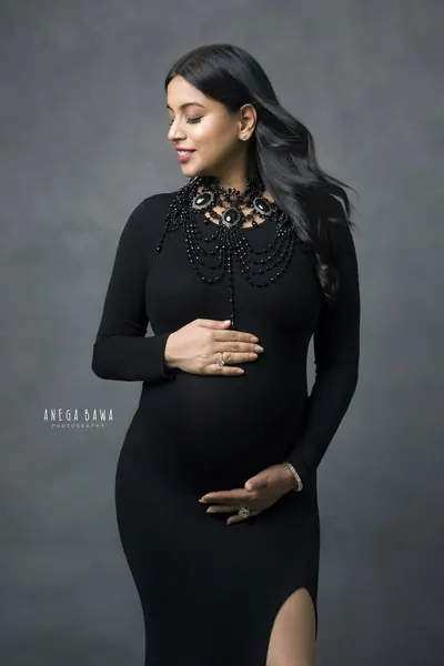 Expecting mother in a black long-sleeve fitted gown with a jewelled beaded bib necklace and a thigh slit, looking down, hands on her bump