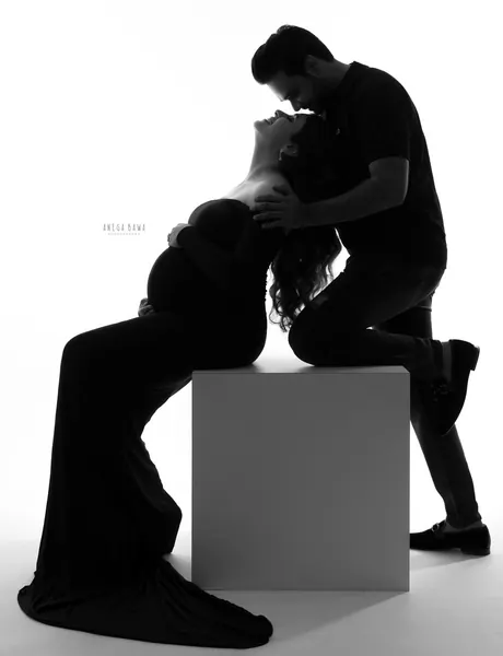 High-key black silhouette of a couple, wife leaning back seated on a white block in a long black gown with her head tilted up while her