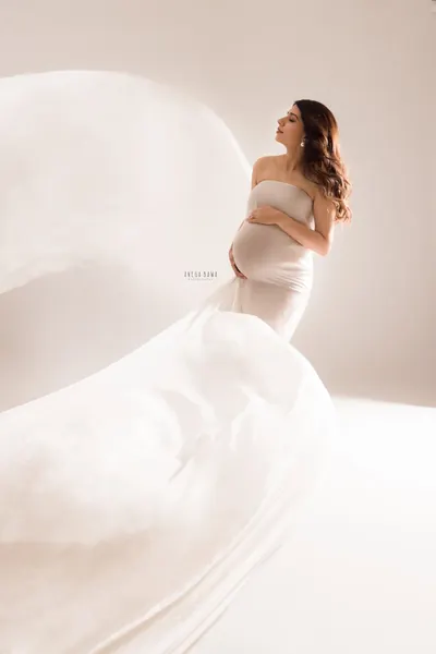 Expecting mother in a cream strapless fitted gown with huge billowing cream drapes sweeping across the frame, looking up, hand on her bump
