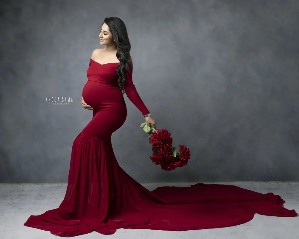 Expecting mother in a deep red off-shoulder long-sleeve mermaid gown with a long train spread on the floor, holding a red rose bunch low in