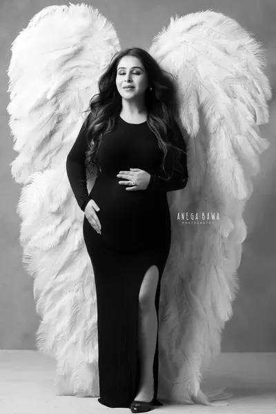 Black and white portrait of expecting mother in a black long-sleeve fitted gown with a high slit standing in front of large white feather