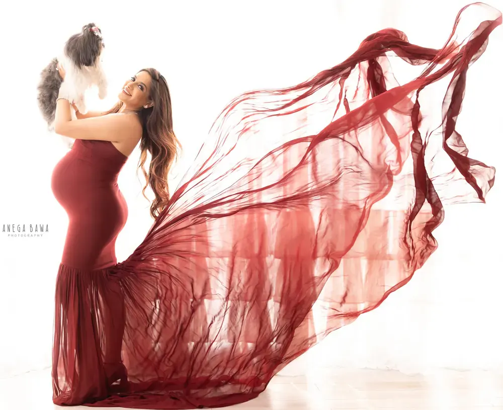 Expecting mother in a red strapless fitted gown with a long sheer red train flying behind her, holding a small white dog up in her arms